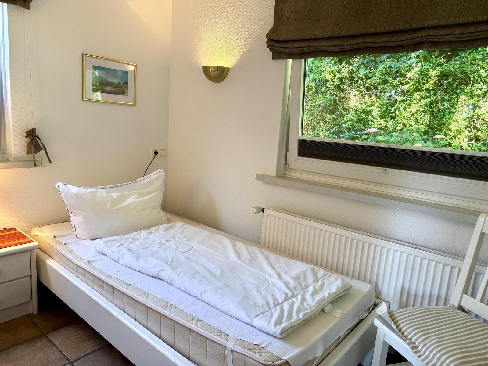 Ein Schlafzimmer mit Bett, Fenster und Stuhl. Draußen sind Bäume sichtbar.