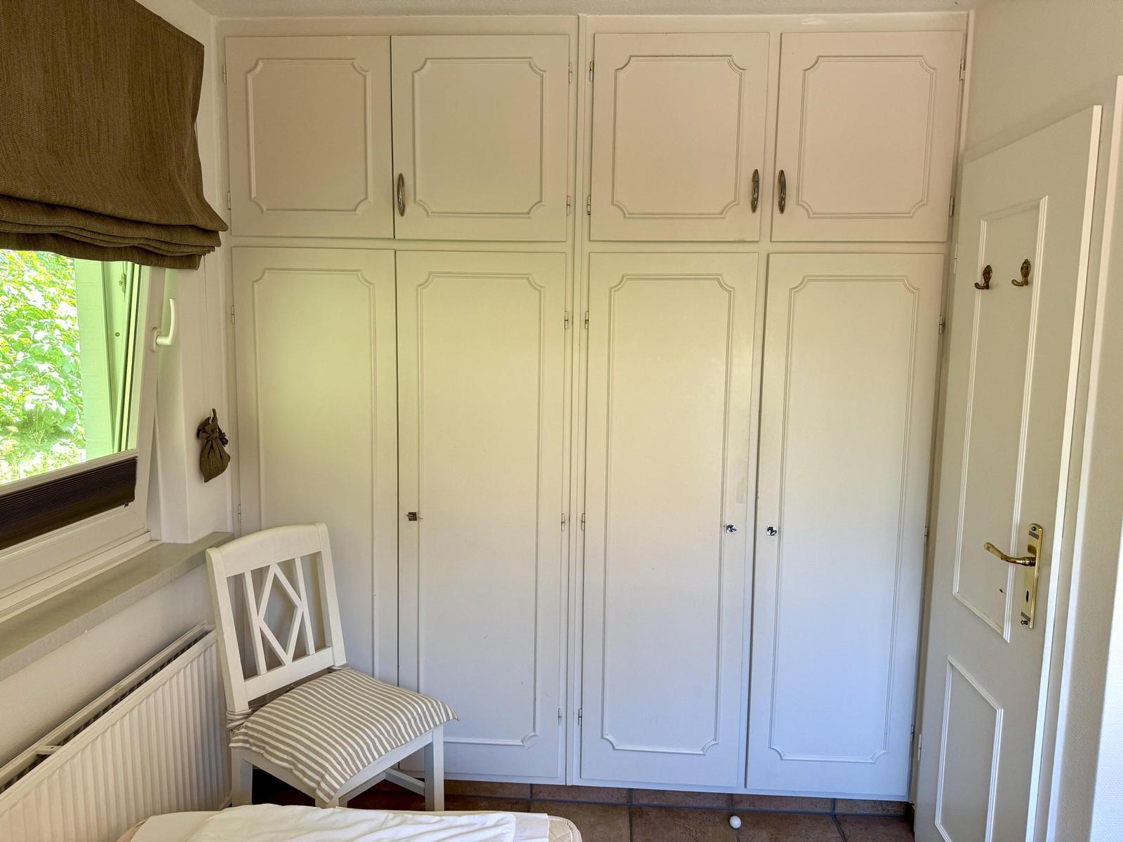 Kleines Zimmer mit weißem Schrank, Stuhl und Fenster mit Blick auf Grün.