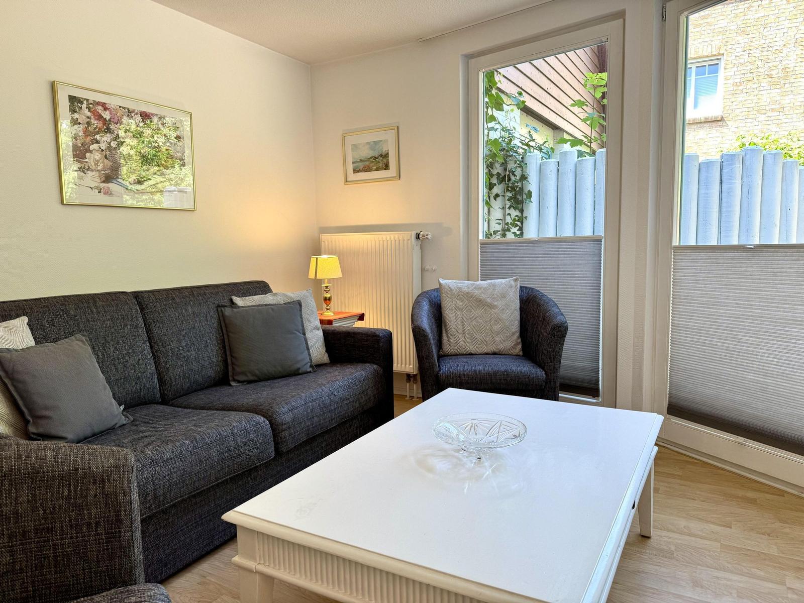 Wohnzimmer mit Sofa, Sessel, Tisch und Fenster zur Außenterrasse.