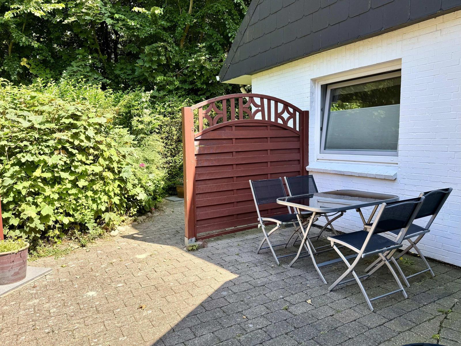 Terrasse mit Tisch und Stühlen neben weißer Hauswand und rotem Tor.
