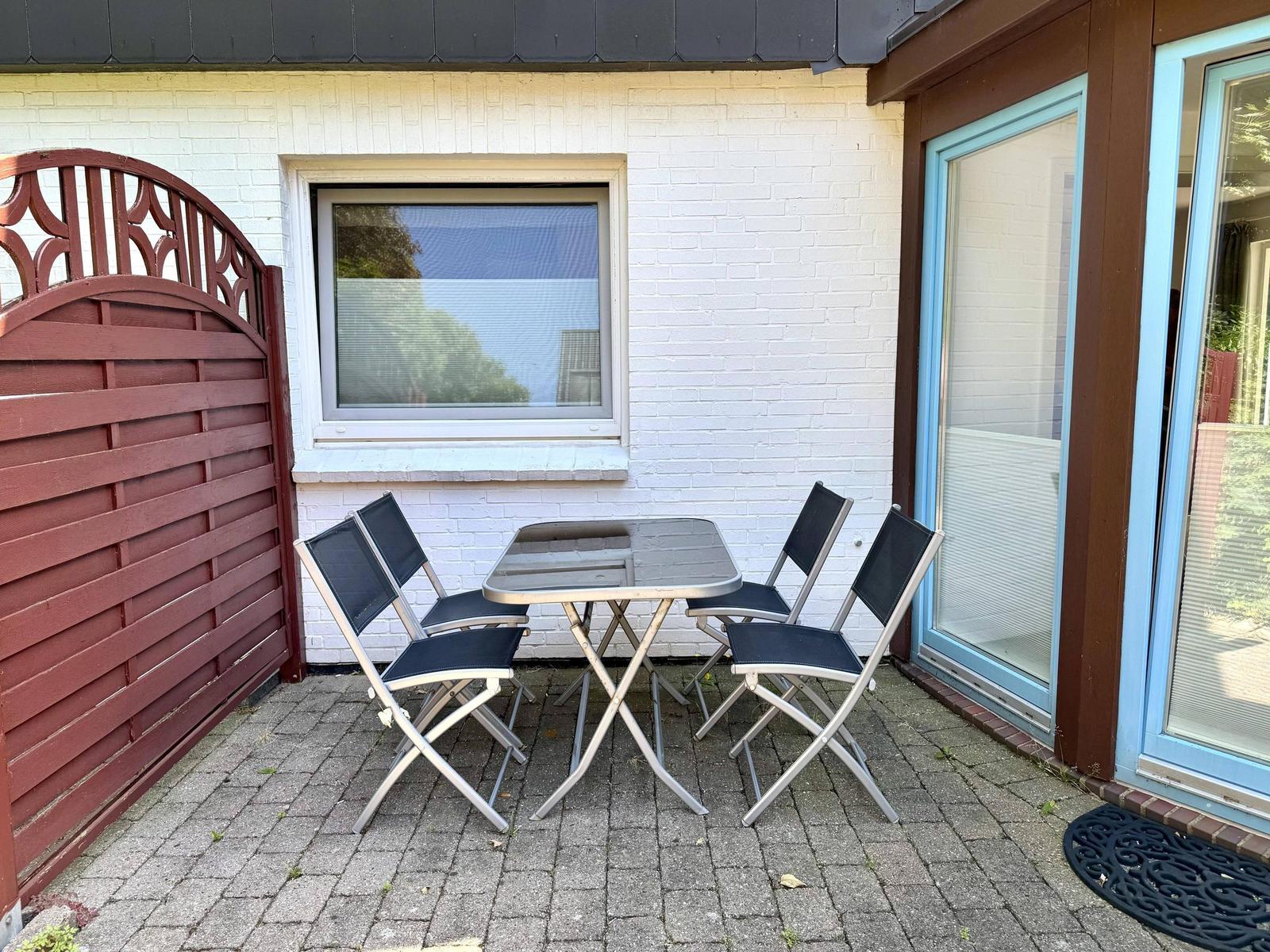 Terrasse mit Tisch und vier Stühlen vor weißem Haus mit Fenster und Glastür.