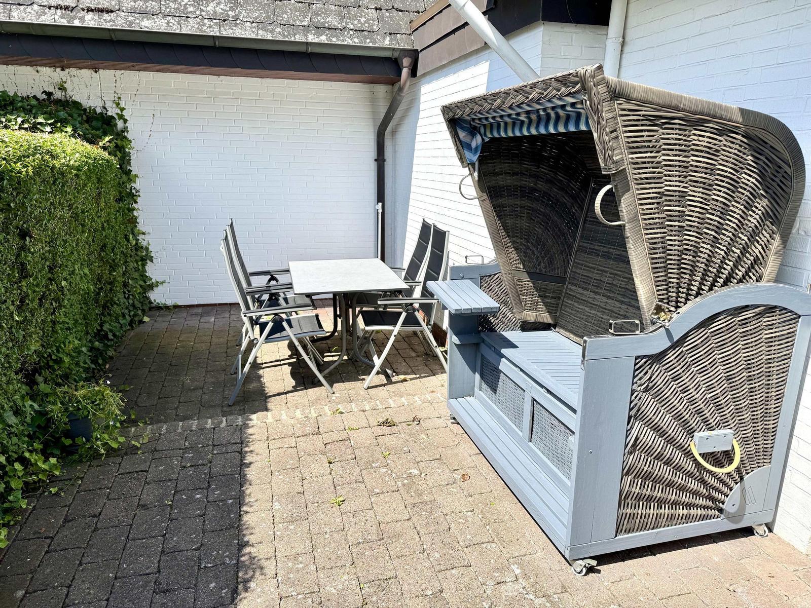 Terrasse mit Tisch, Stühlen und Strandkorb. Weiße Wand und Hecke im Hintergrund.