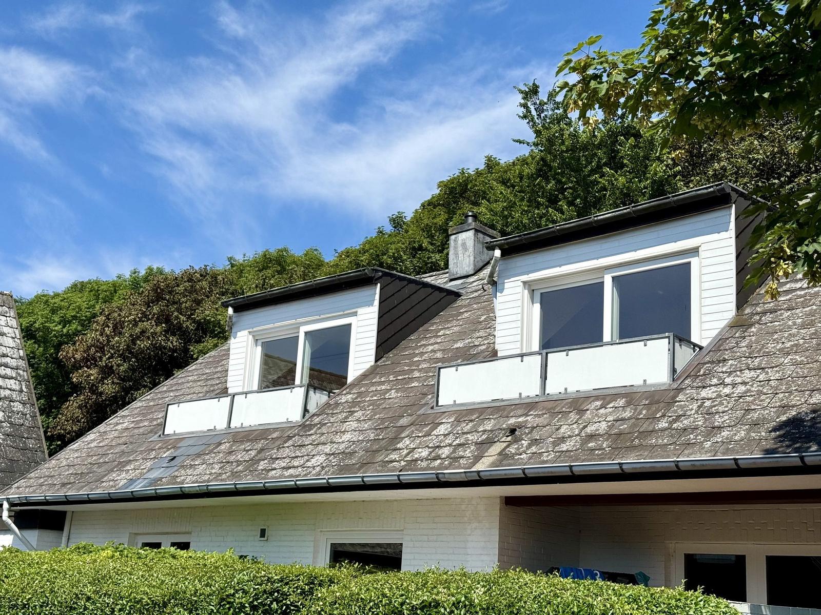 House with dormer windows and balcony, surrounded by trees under blue sky.