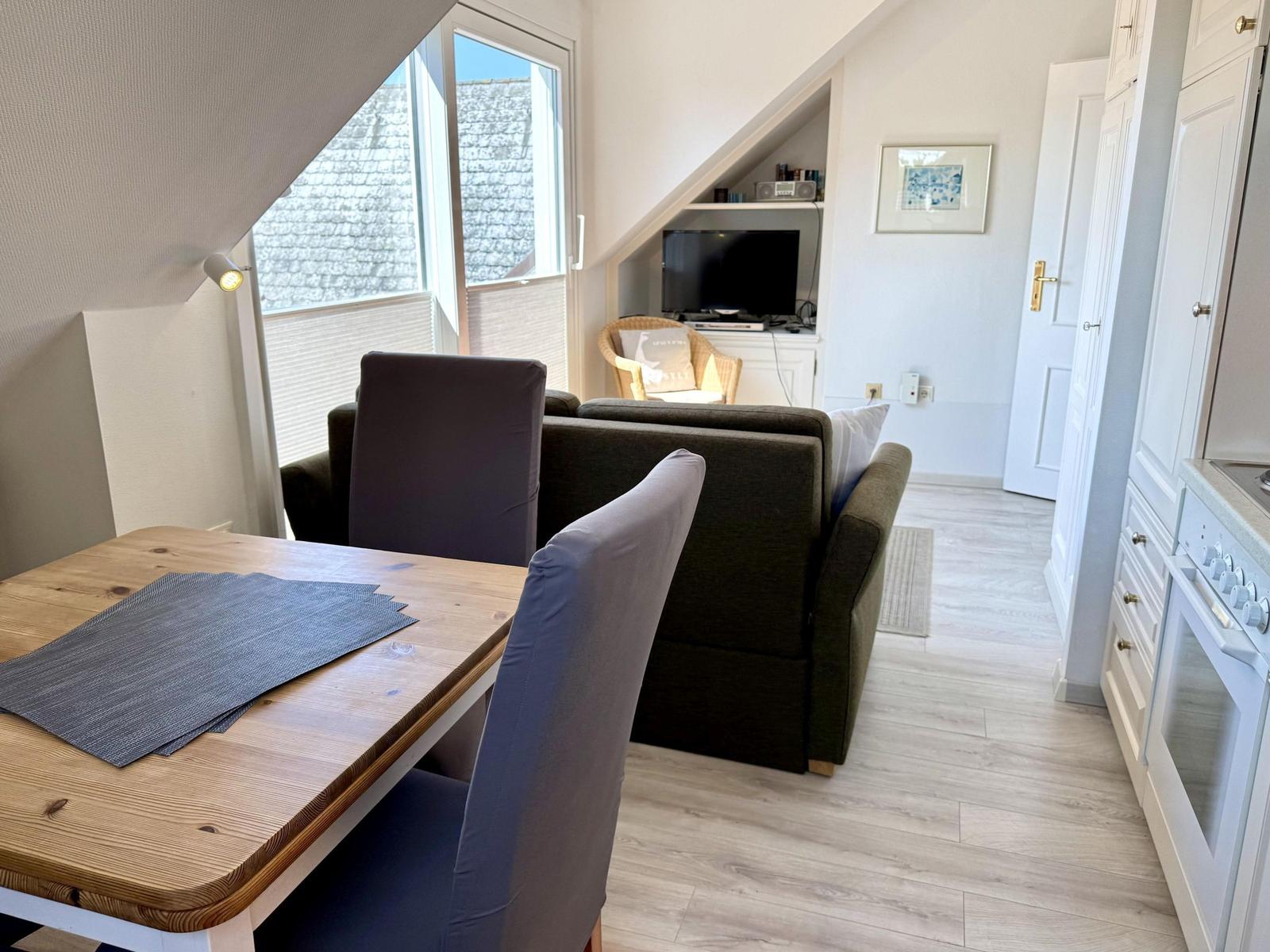 Dining area with table and chairs, living area with sofa and TV, kitchen with white cabinets.