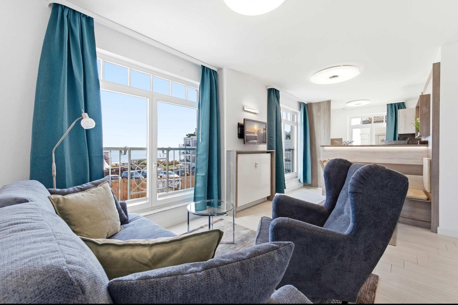 Wohnzimmer mit Sofa, Sessel und Blick auf Balkon. Teleskoplampe und Fernseher an Wand.