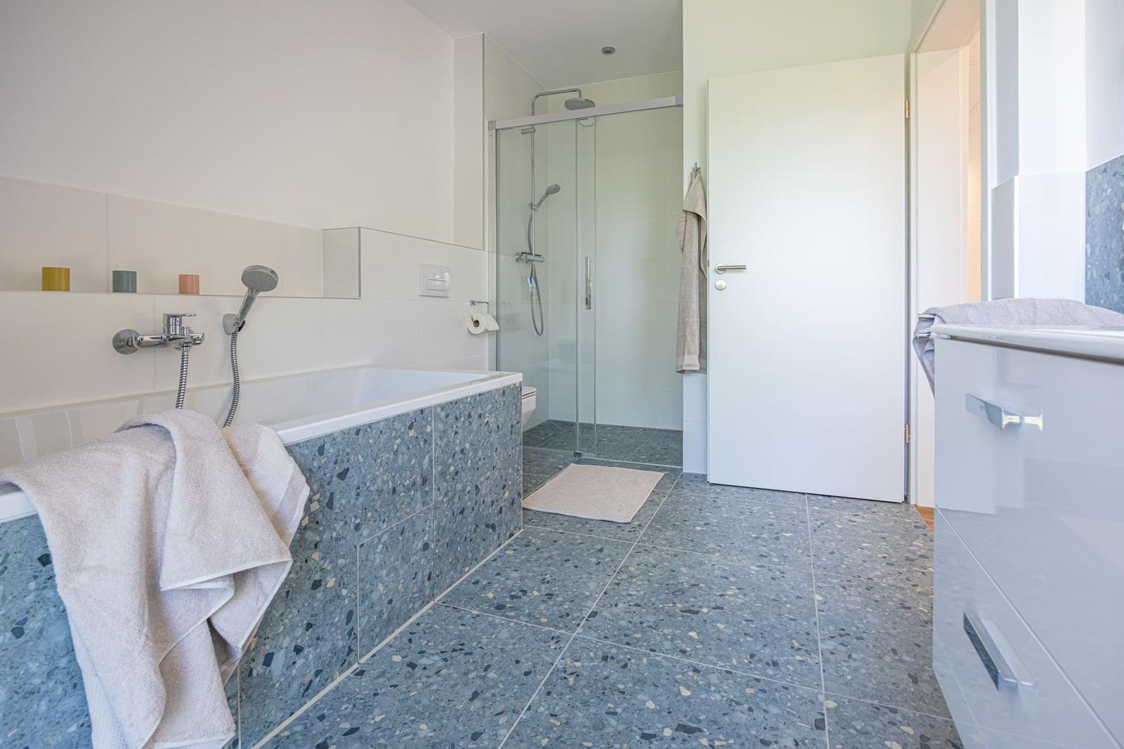 Badezimmer mit Badewanne, Dusche und Waschtisch. Boden und Wandfliesen sind terrazzoartig.