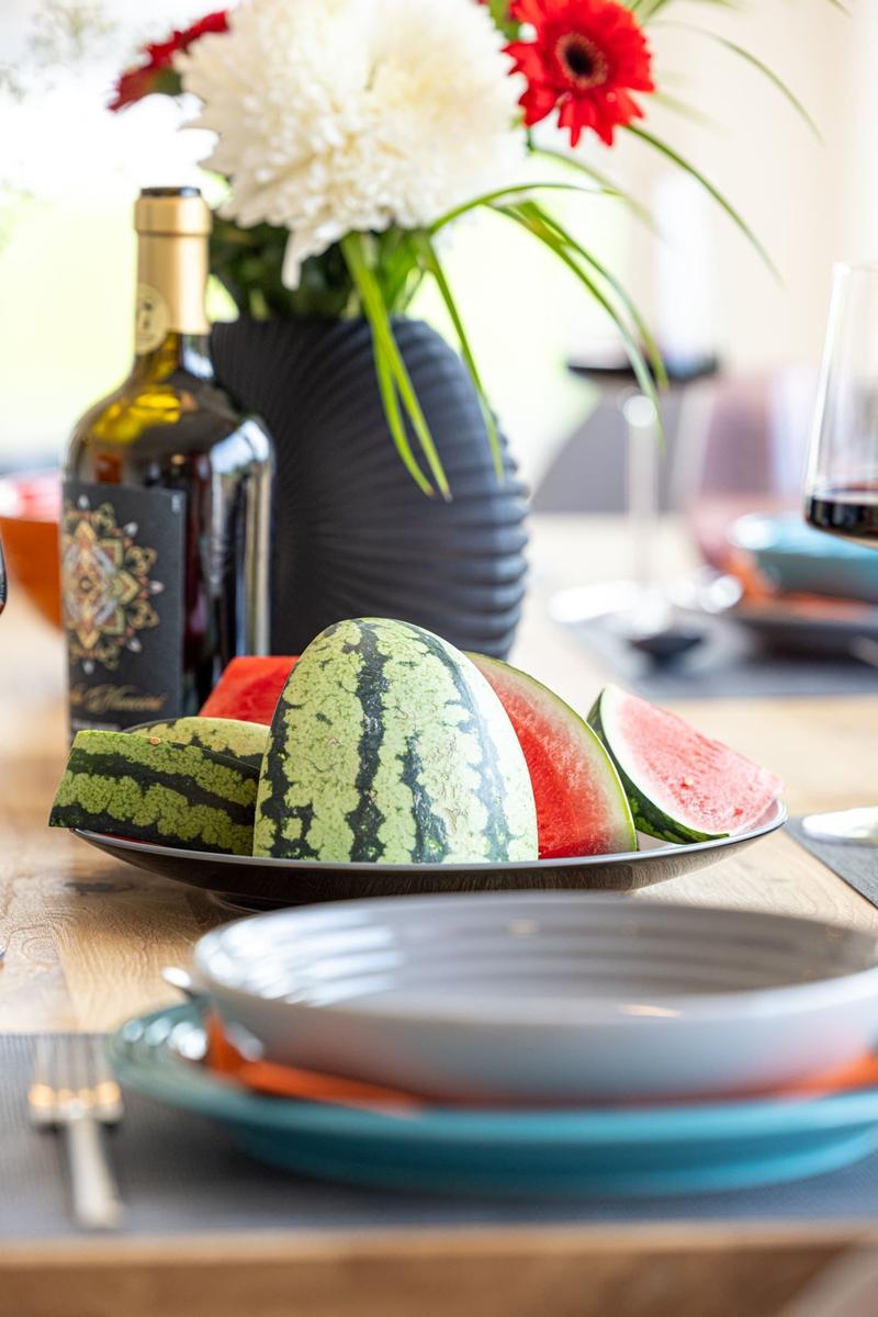 Tisch mit Wassermelonen, Wein und Blumenarrangement.