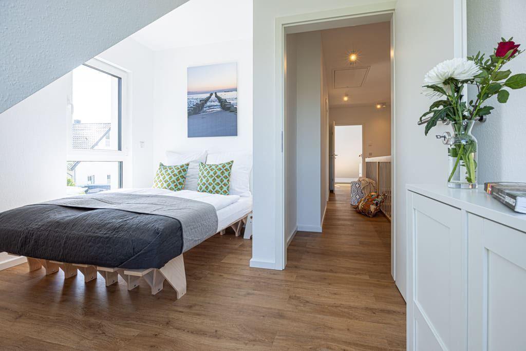 Schlafzimmer mit Bett, Fenster und Bild an der Wand. Blick in den Flur mit Holzboden.