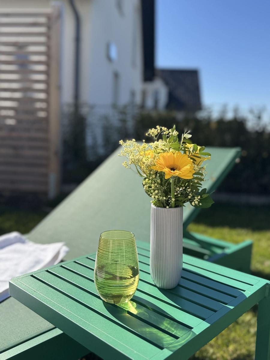 Auf einem grünen Tisch steht ein Glas und eine Vase mit Blumen. Im Hintergrund liegt eine Liege.