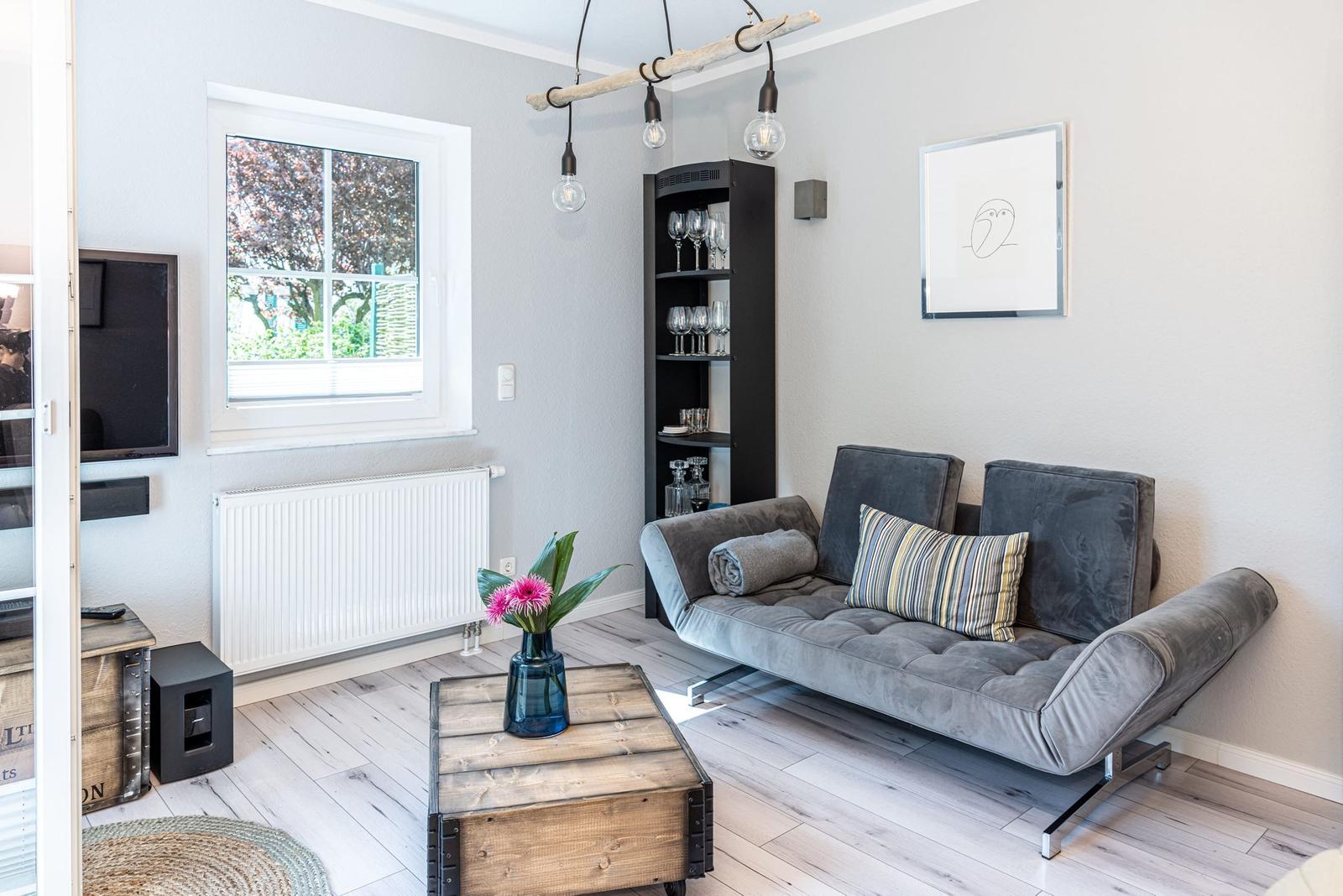 Ein Wohnzimmer mit grauem Sofa, Holzboden und Fenster mit Blick auf Bäume.