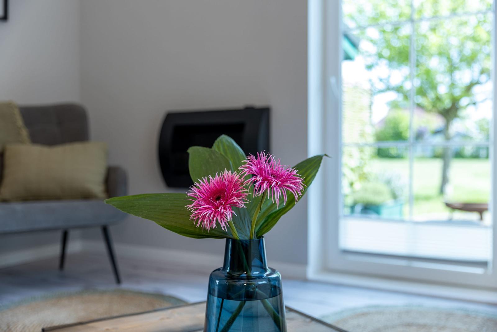 Ein blauer Vasen mit zwei pinken Blumen auf einem Tisch. Im Hintergrund ein Sofa und ein Fenster.