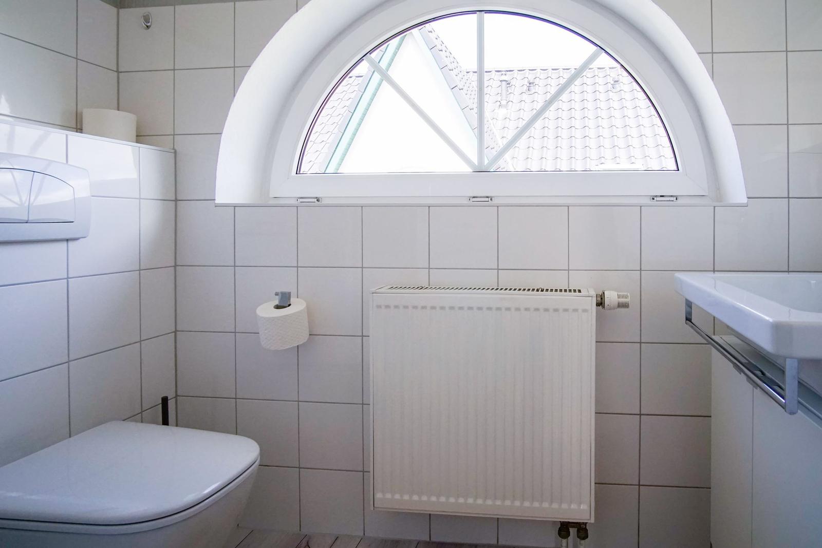Badezimmer mit Toilette, Waschbecken und Heizkörper unter halbrundem Fenster.