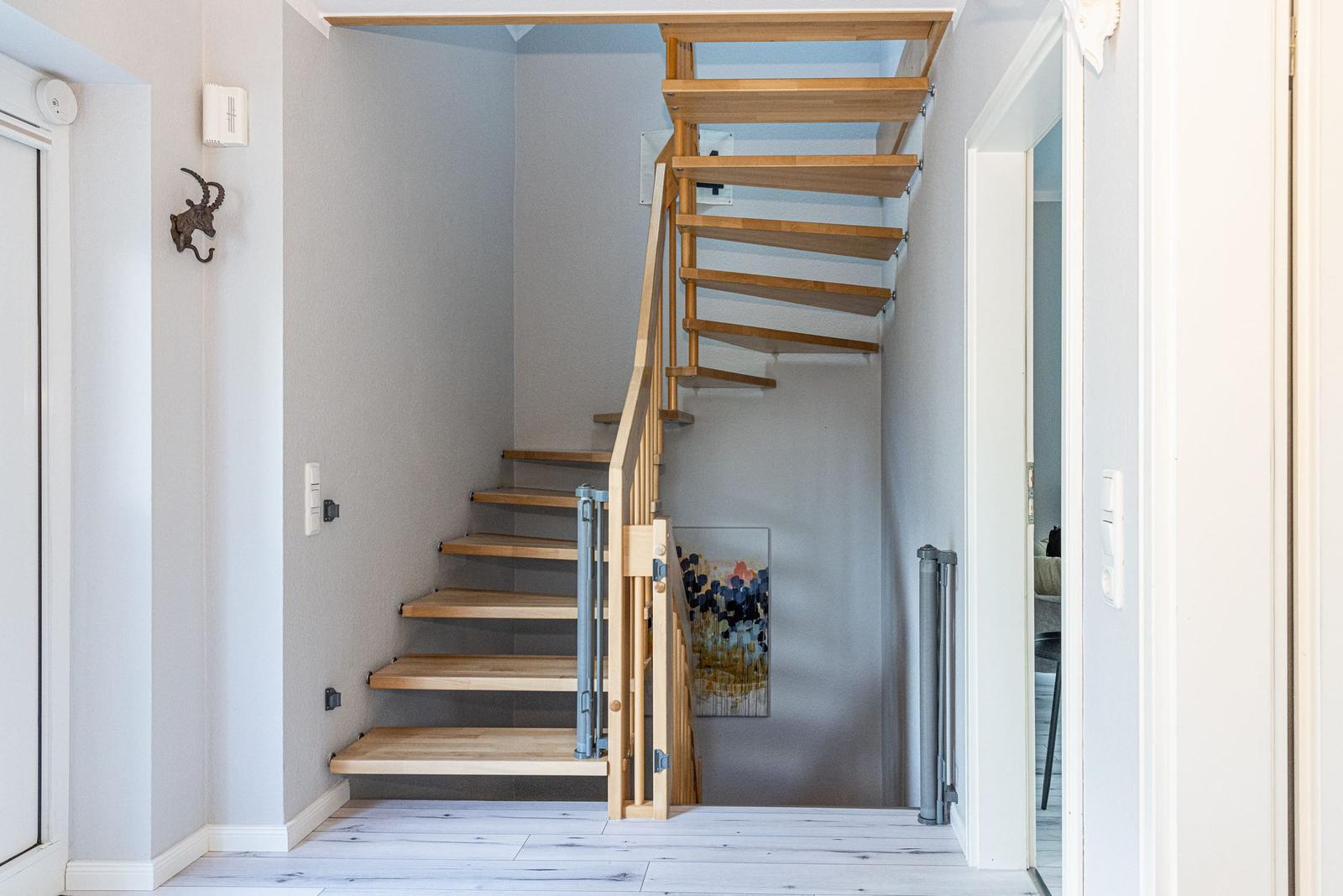 Holztreppe mit Geländer führt nach oben. Wandbild und Türöffnung sind sichtbar.