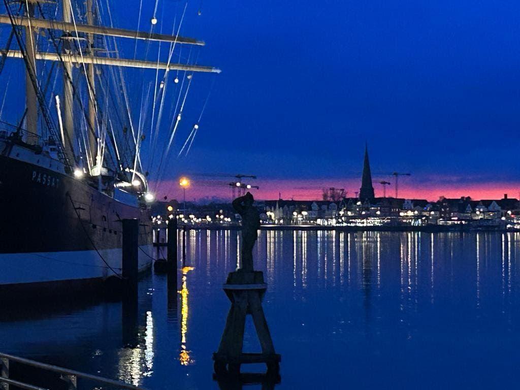 Historisches Schiff und Statue am Hafen bei Dämmerung mit beleuchteter Stadt im Hintergrund.