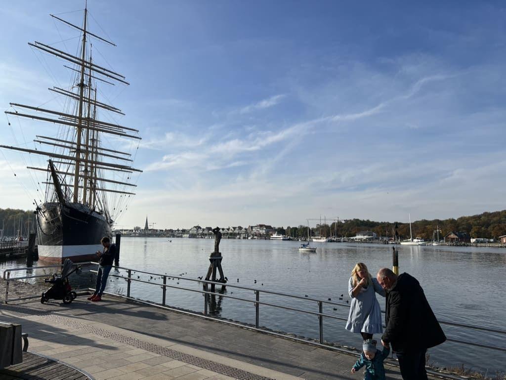 Hafen mit Segelschiff, Promenade und Besuchern an der Wasserkante.