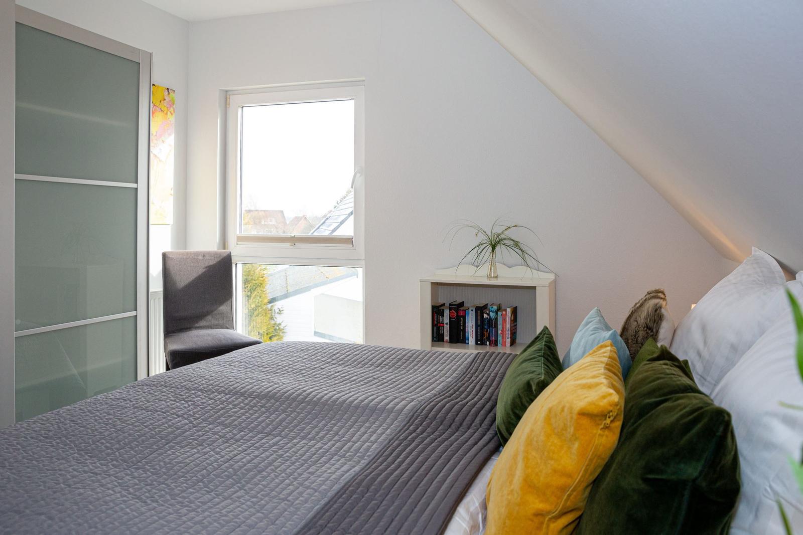 Schlafzimmer mit Bett, Fenster, Stuhl und Bücherregal unter Dachschrägen.