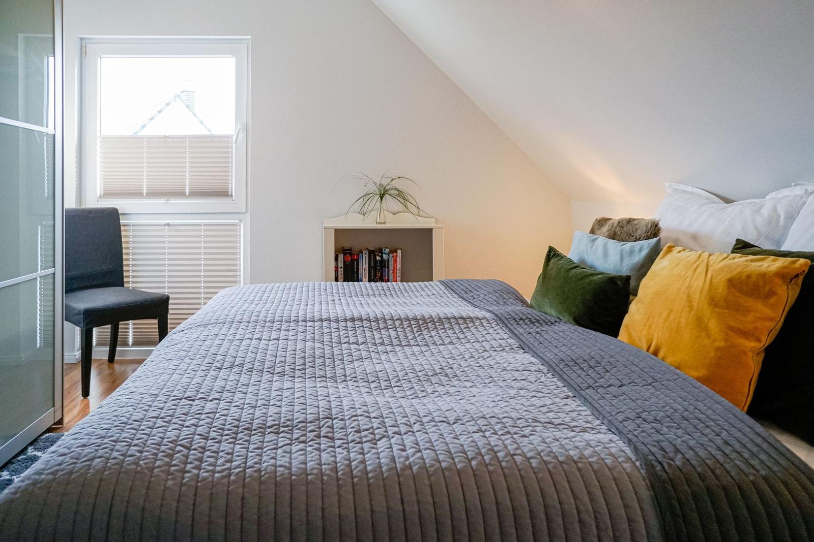 Schlafzimmer mit Dachschräge, Bett mit Kissen und kleinem Bücherregal.