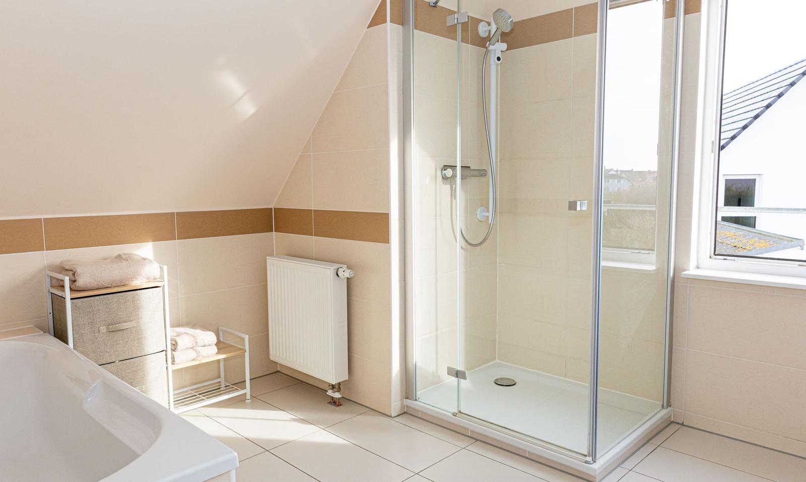 Badezimmer mit Dusche, Badewanne und Fenster. Holzregale und Heizkörper sichtbar.