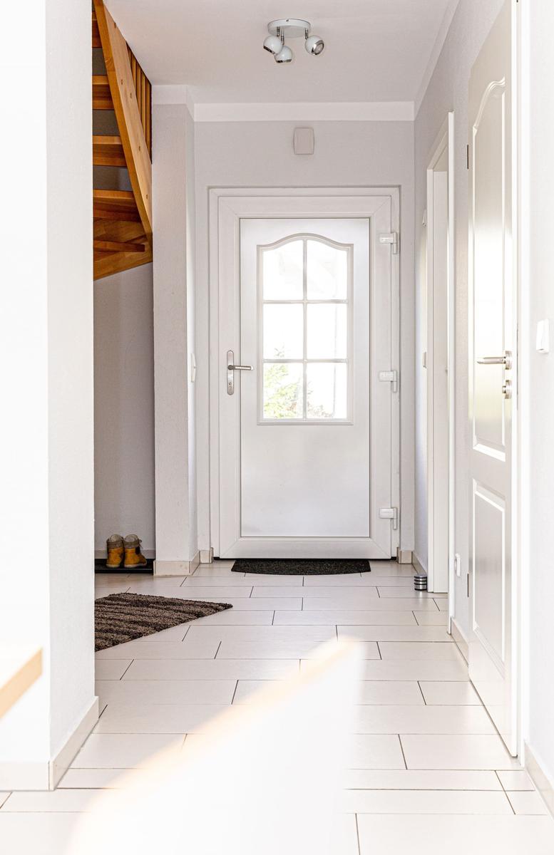 Hausflur mit weißen Wänden, Fliesenboden und Holztreppe. Eingangstür mit Glasfenster.