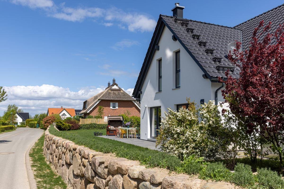 Modernes Haus mit Terrasse, Garten und Steinmauer. Hintergrund zeigt traditionelle Gebäude.