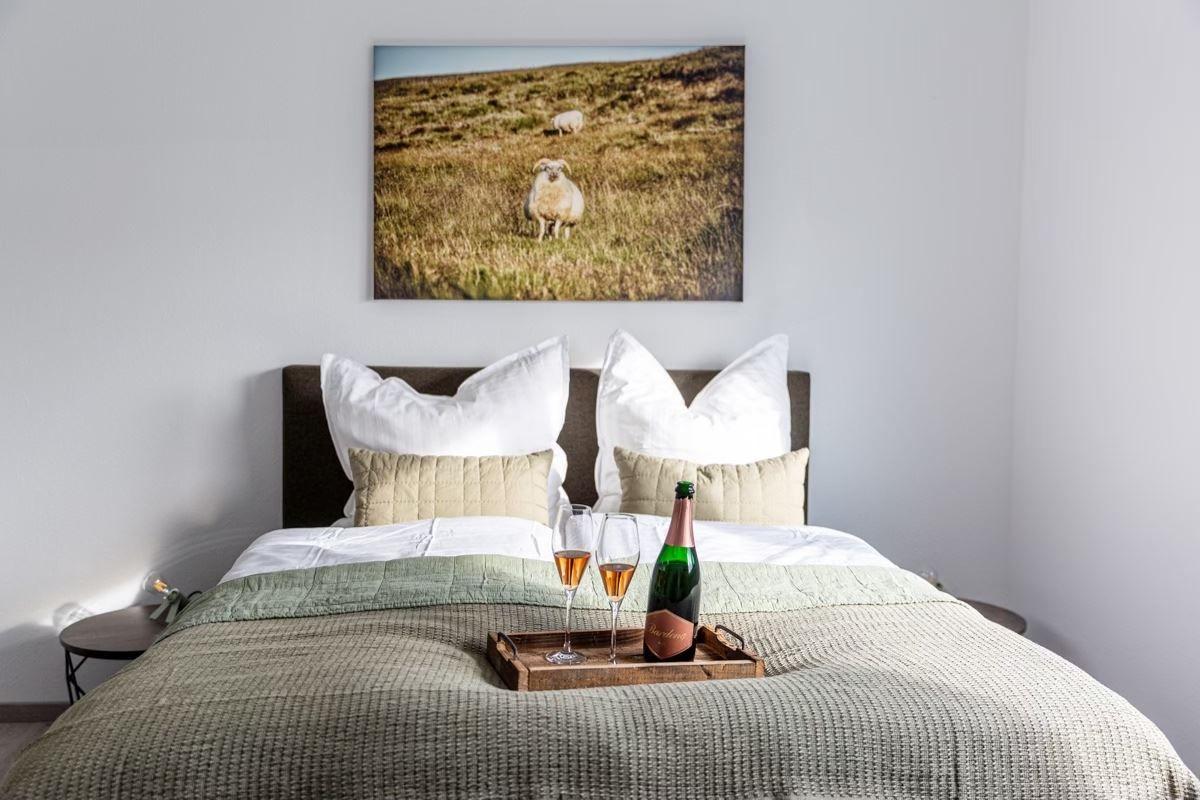 Ein Bett mit Kissen und einem Holztablett mit Champagner und Gläsern. Über dem Bett hängt ein Bild von Schafen.