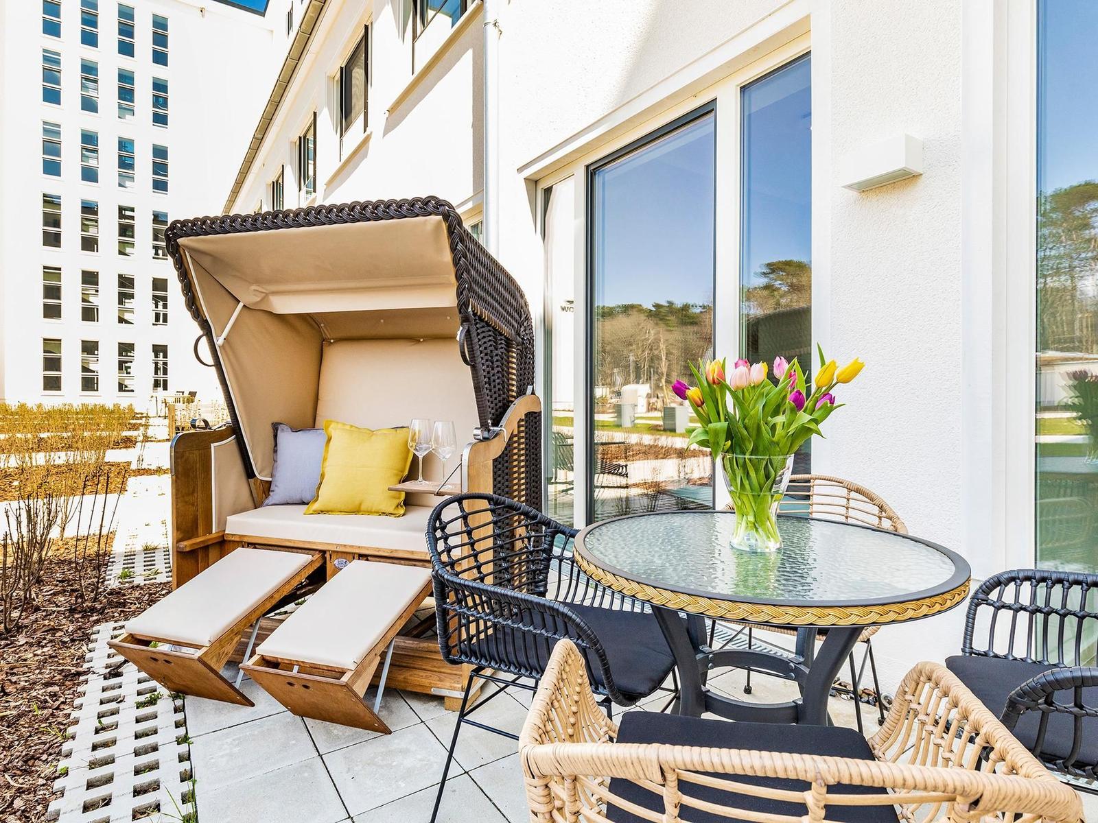Terrasse mit Liege, Tisch und Stühlen. Blumen im Glas auf Tisch. Hintergrund: Gebäude mit Fenstern.