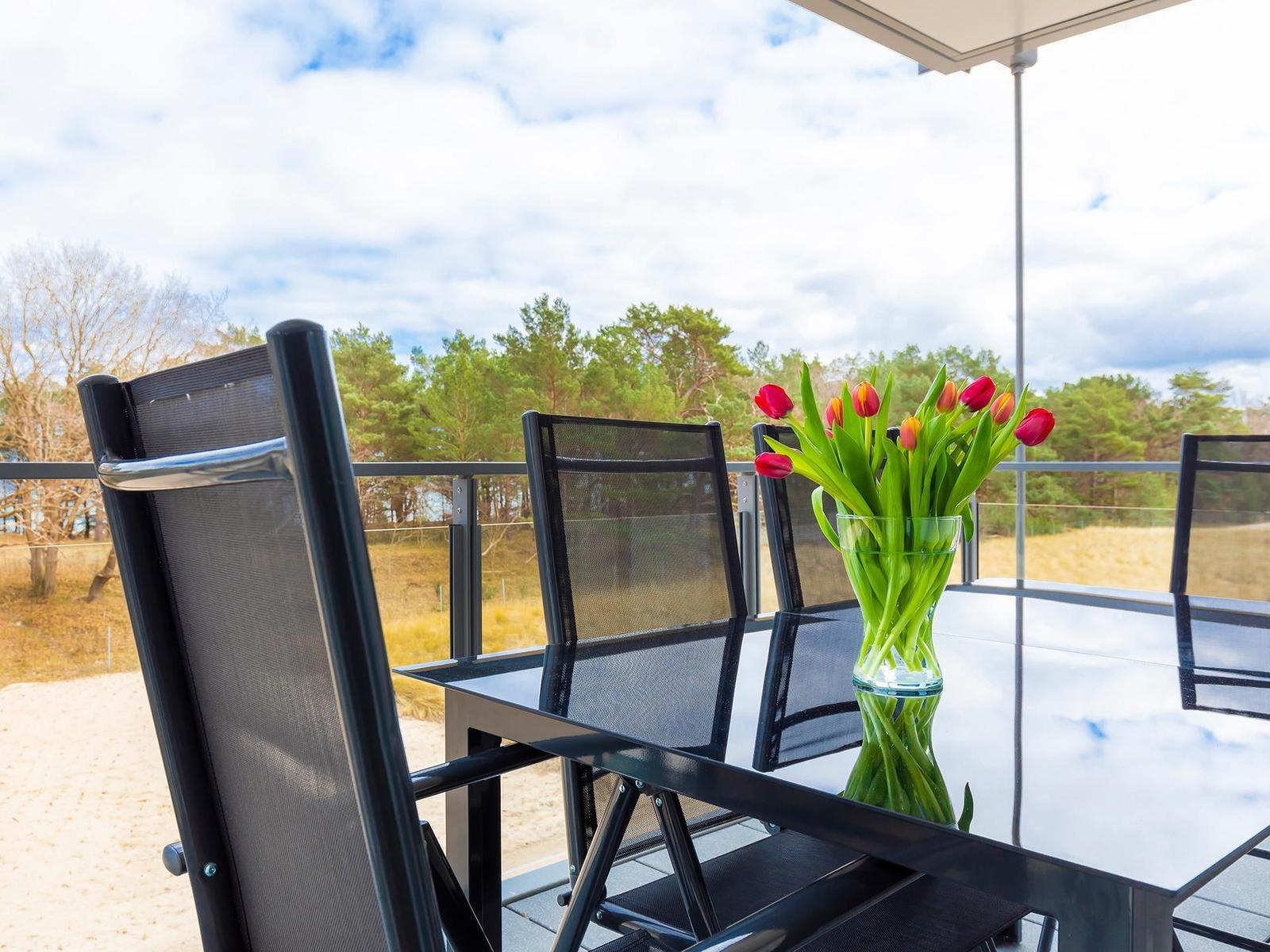 Terrasse mit Tisch, Stühlen und Tulpenblumen. Blick auf Wald und Himmel.