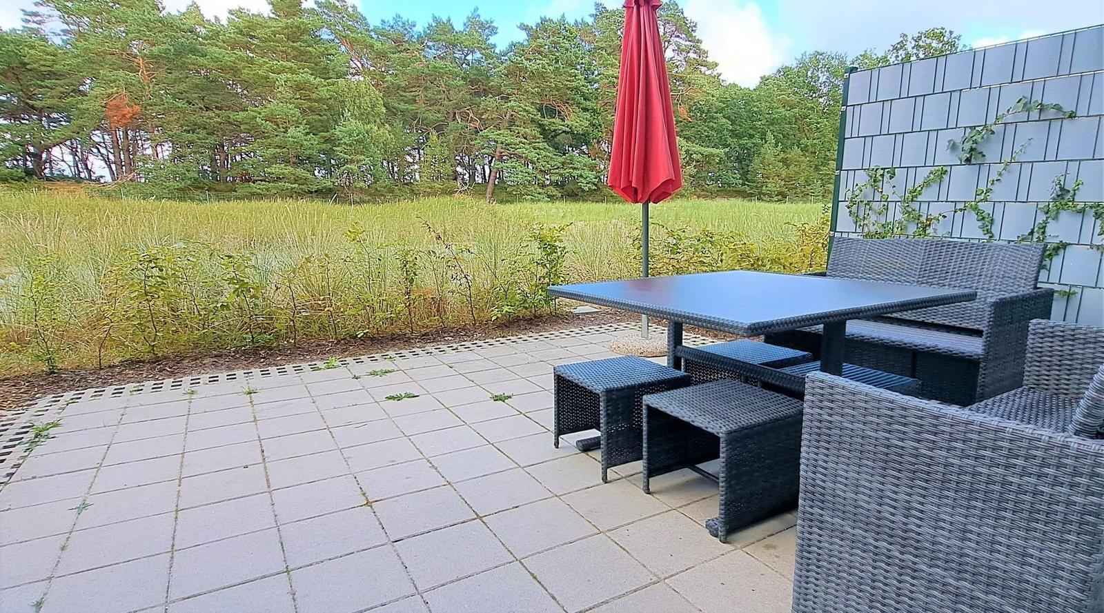 Terrasse mit Tisch, Stühlen und einem roten Sonnenschirm. Hintergrund: Grasland und Wald.