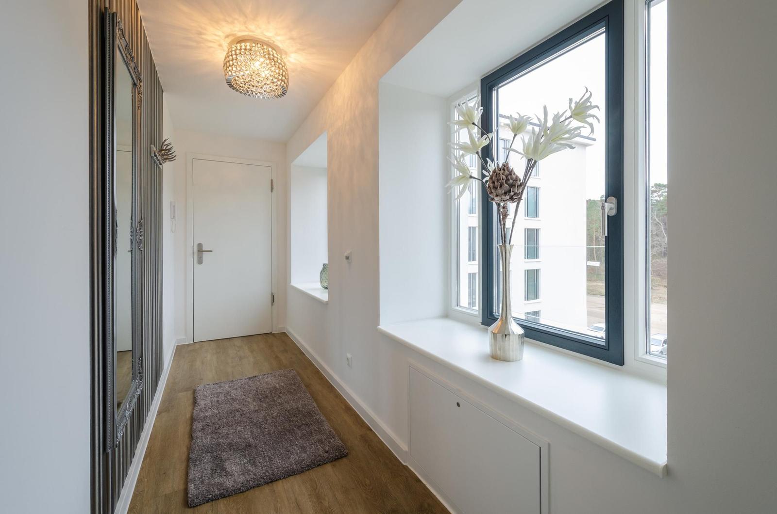 Hallway with wooden floor, mirror, window, and rug. Ceiling light and flowers on windowsill.