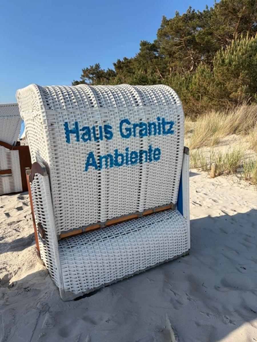 White beach chair with inscription Haus Granitz Ambiente stands on sand before trees.