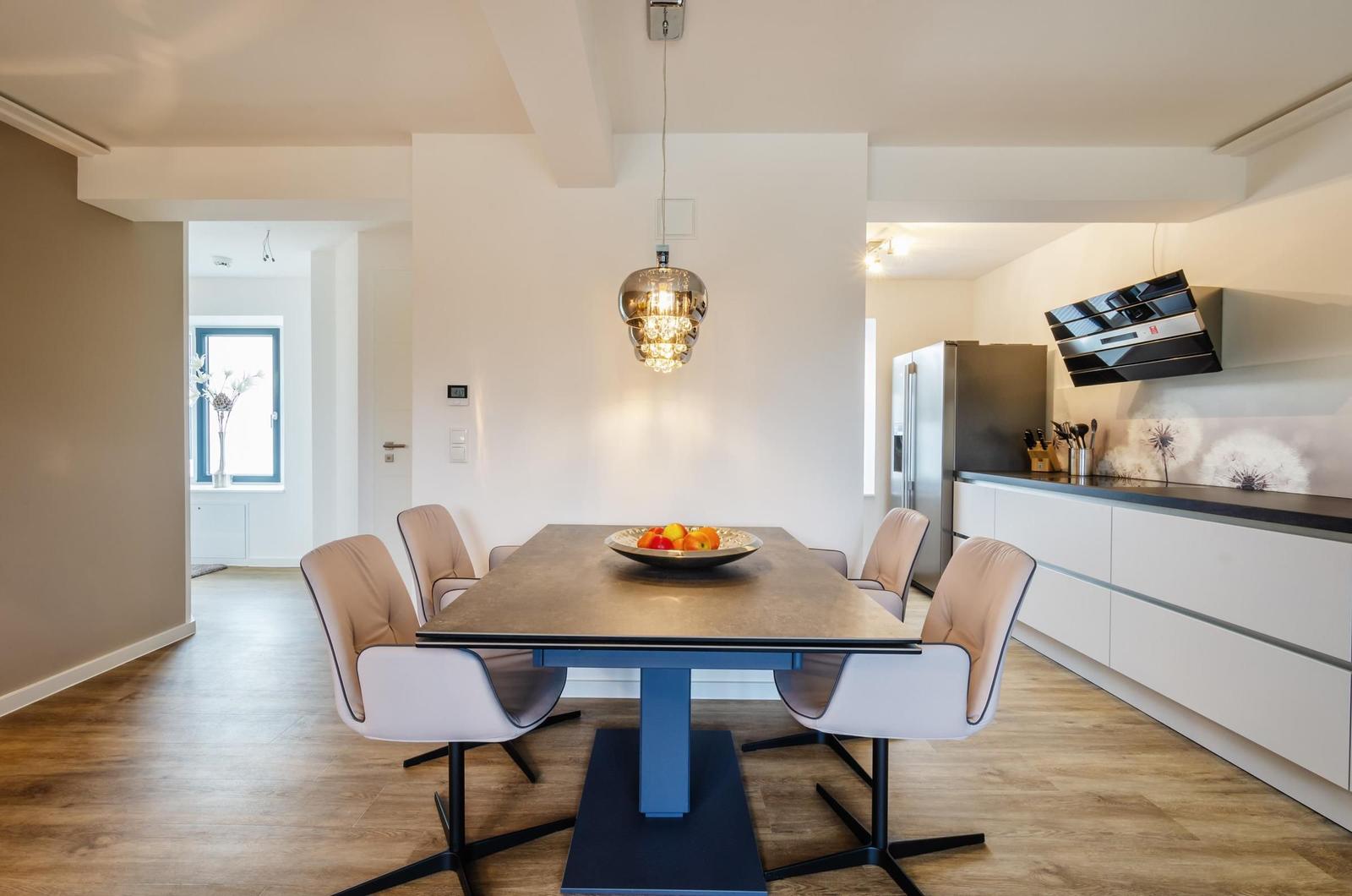 Dining area with table, chairs, and modern lighting. Kitchen with counter and refrigerator.