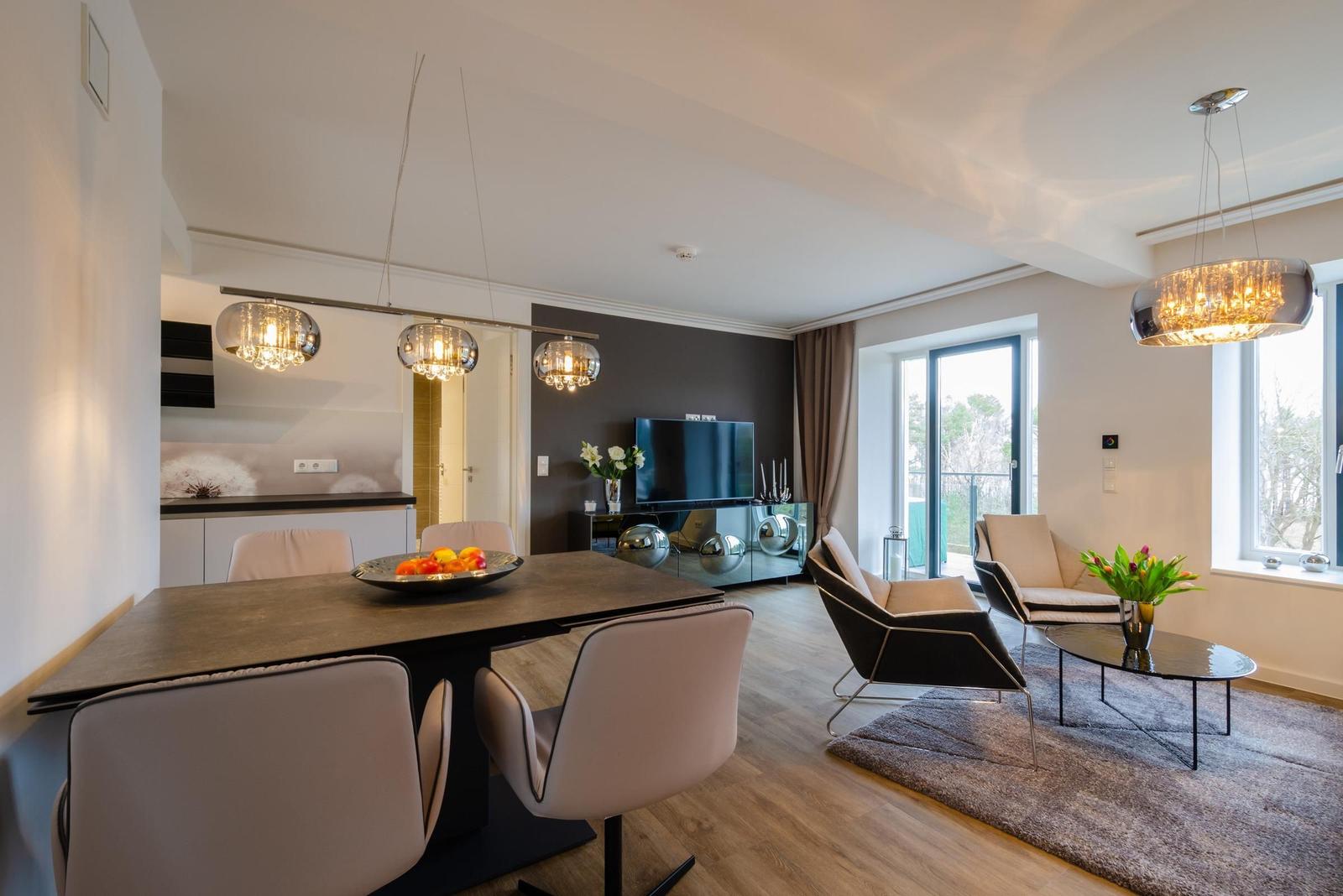 Dining area with table and chairs, living room with armchairs and TV, large windows with balcony view.