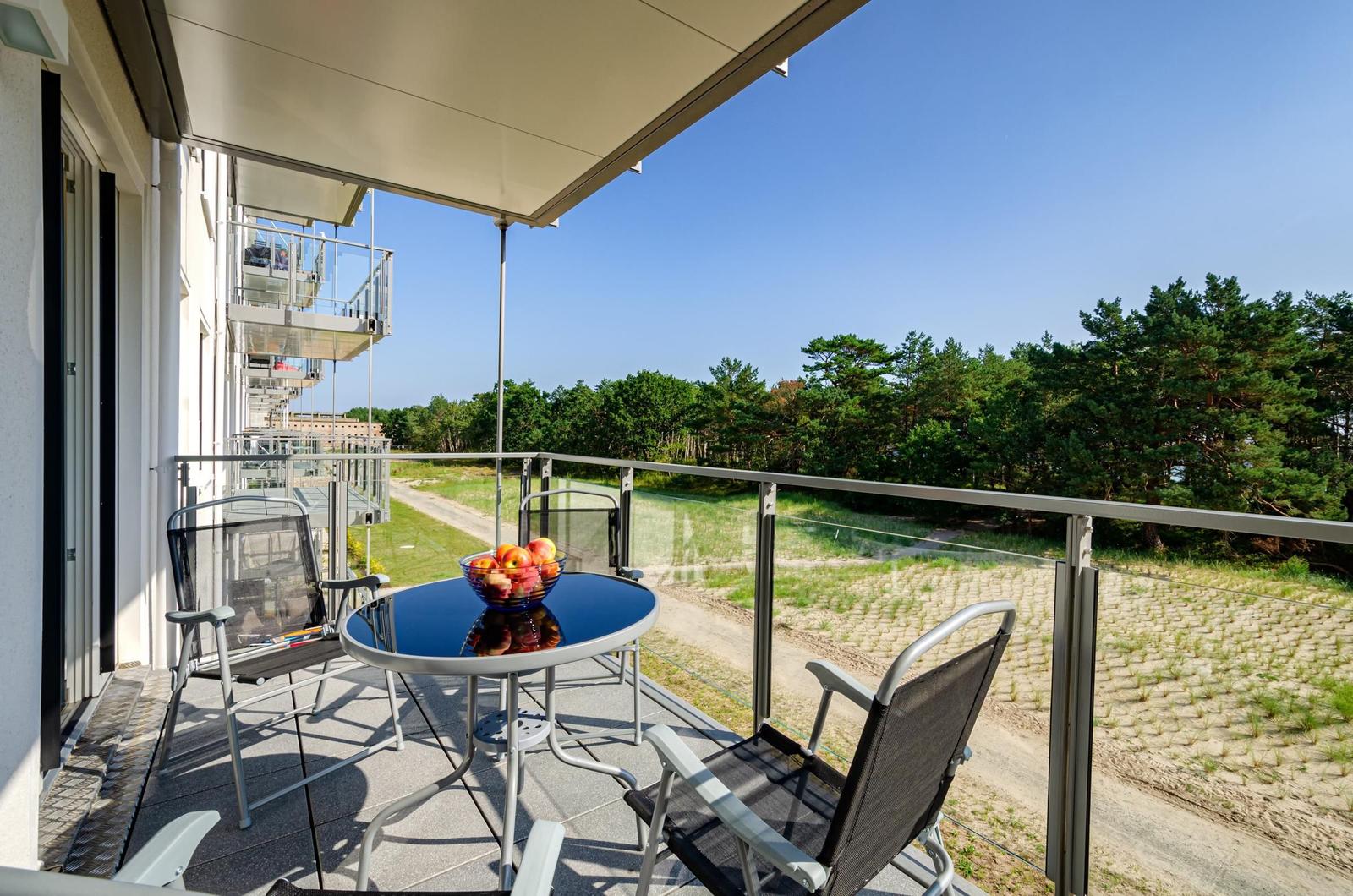 Terrasse mit Tisch, Stühlen und Obstschale. Blick auf Wald und Gebäude.