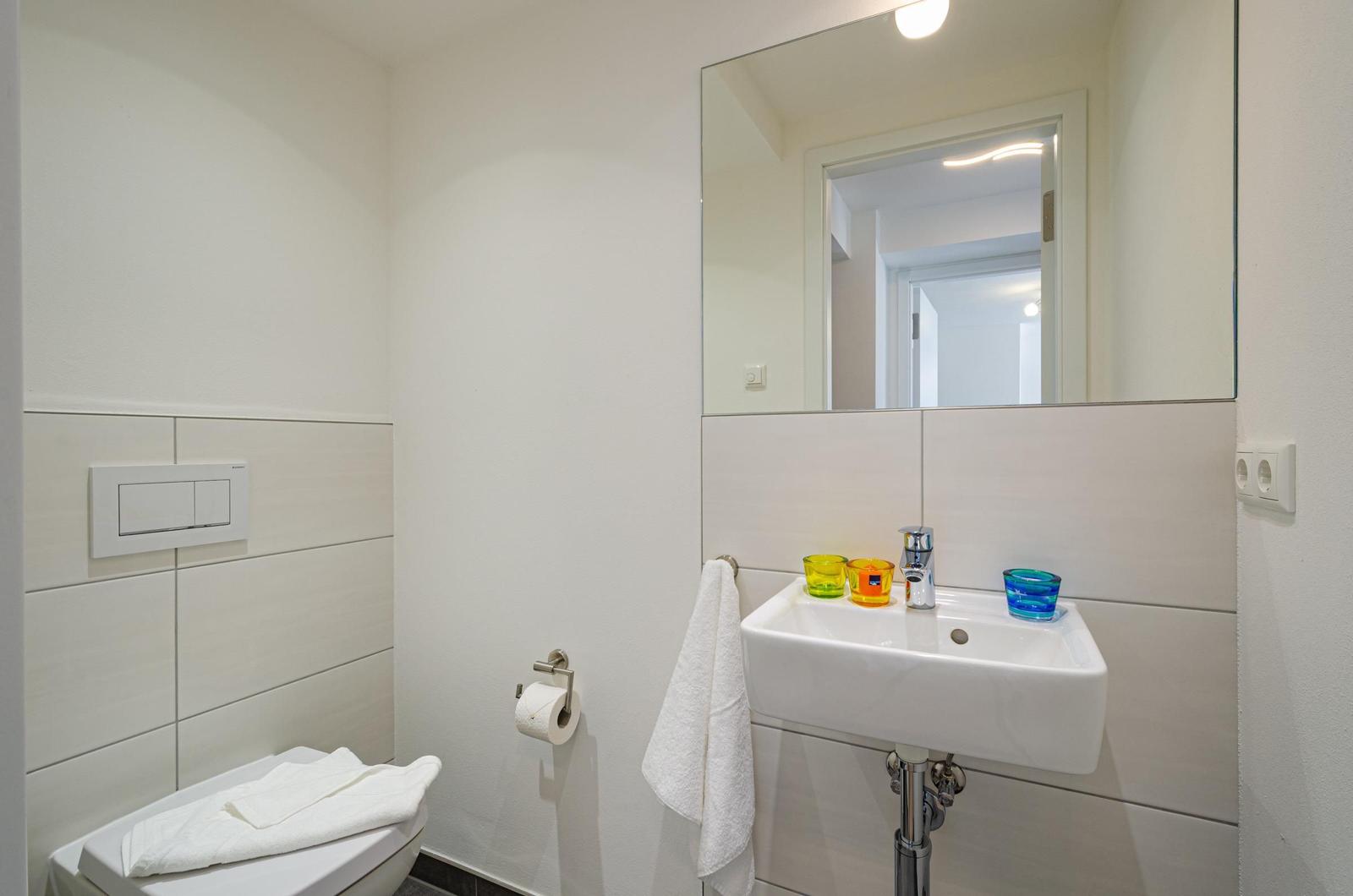 Badezimmer mit Toilette, Waschbecken und Spiegel. Drei farbige Gläser auf dem Waschbecken.