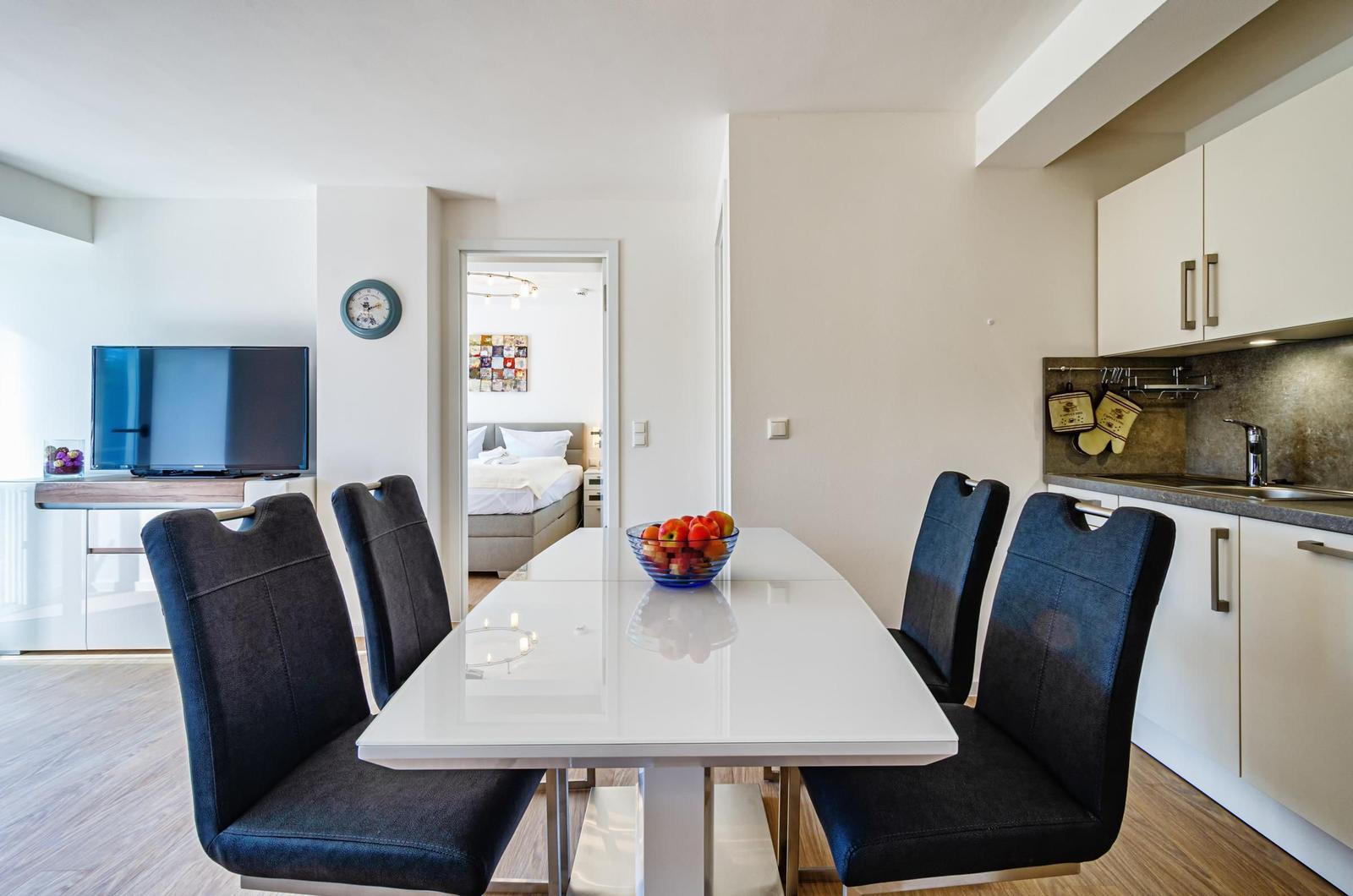 Essbereich mit Tisch und Stühlen, Küche und TV im Hintergrund.