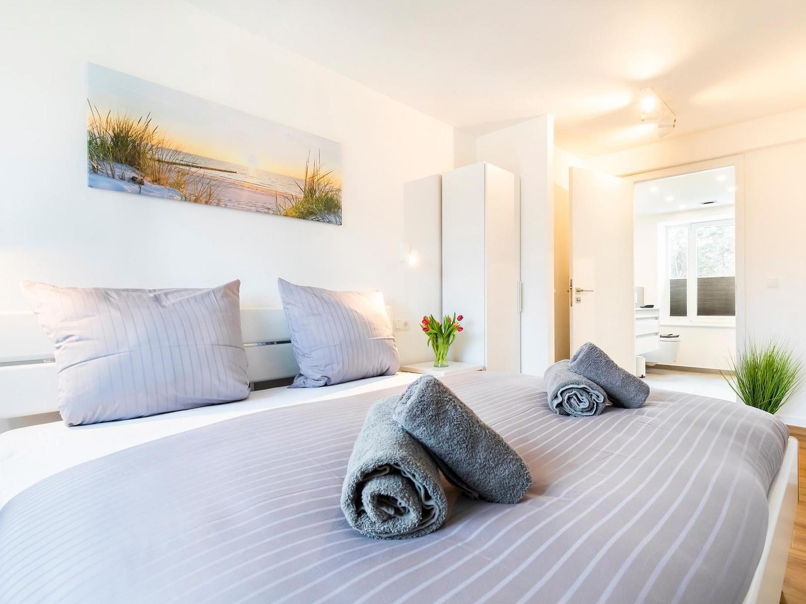 Schlafzimmer mit Bett, Kissen und Handtüchern. Wandbild und Badezimmer sichtbar.