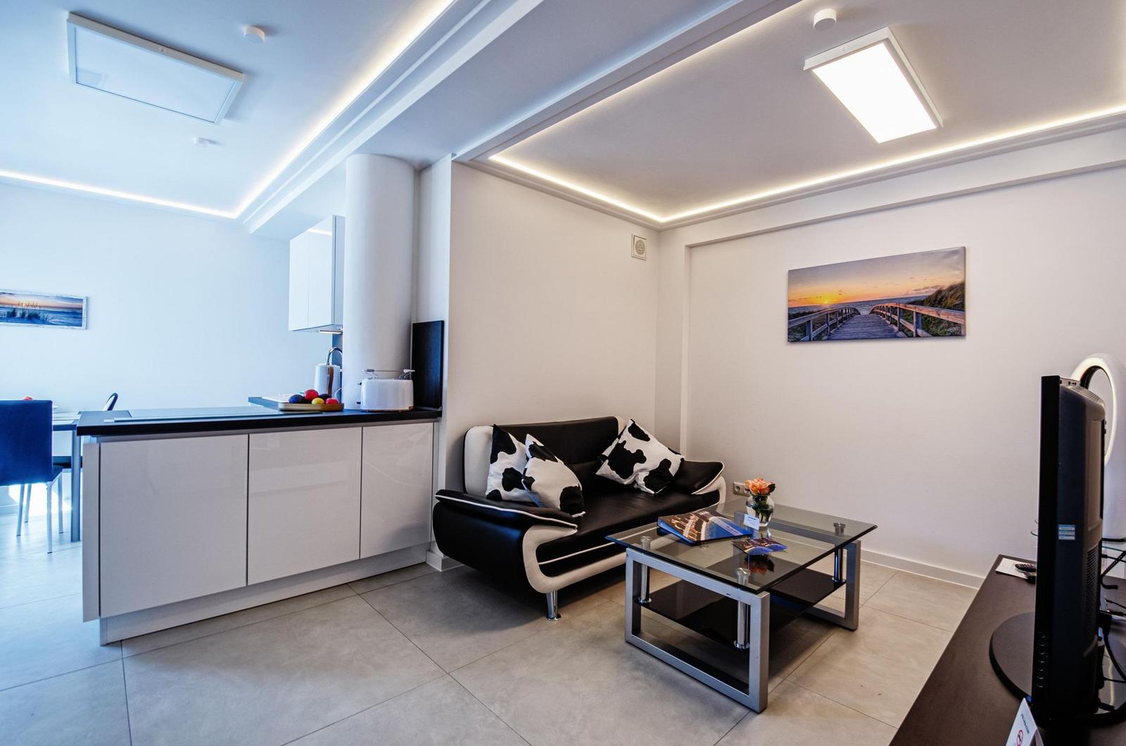 Wohnzimmer mit Sofa, Couchtisch und Fernseher. Küchenbereich mit Arbeitsplatte und Stühlen.