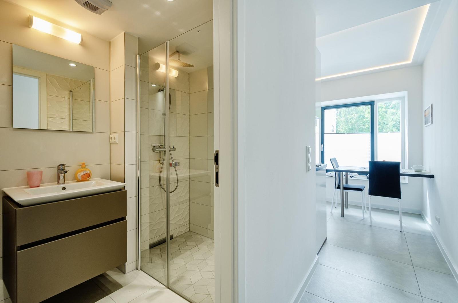 Badezimmer mit Dusche und Waschbecken. Durchgang zu Essbereich mit Tisch und Stühlen.