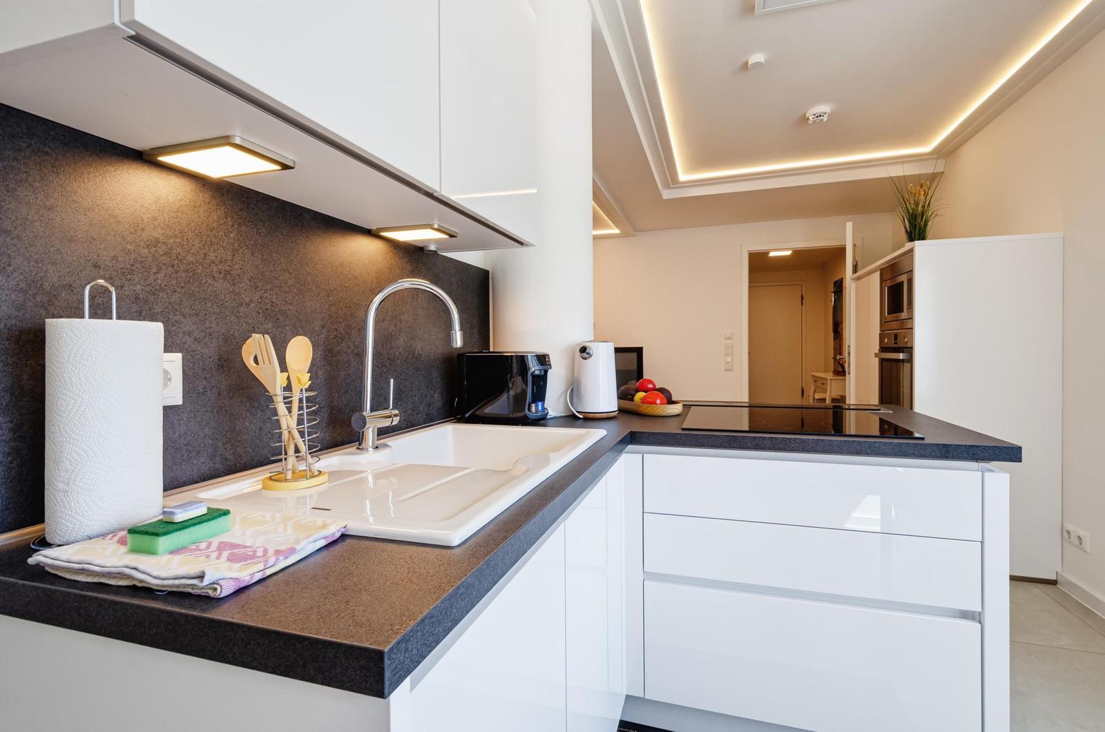 Küche mit weißen Schränken, dunklem Arbeitsplatz und Spüle. Hintergrund zeigt Wohnbereich.