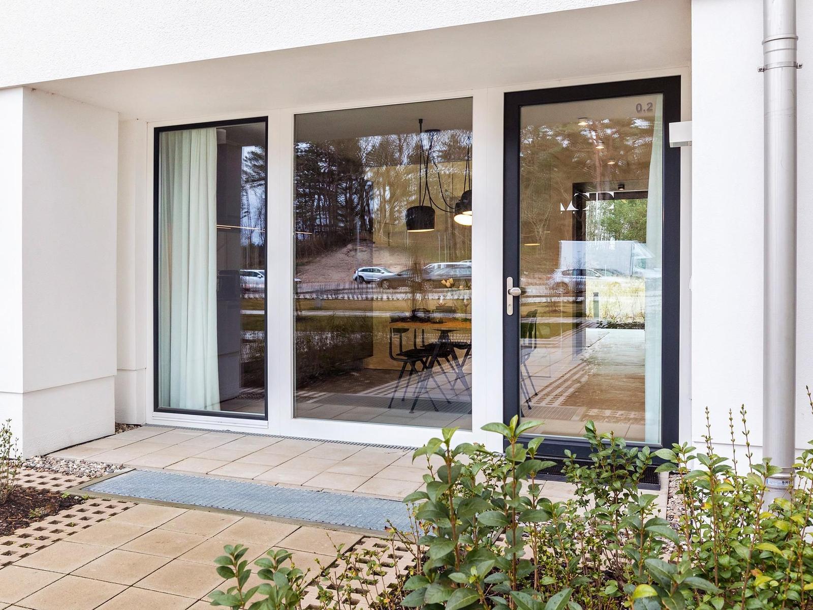 Eingangsbereich mit Glasfront und Terrasse. Im Inneren ist ein Essbereich mit Tisch und Stühlen sichtbar.