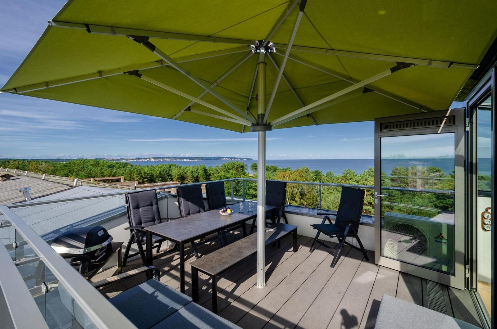 Terrasse mit Sonnenschirm, Tisch, Stühlen und Grill. Blick auf See und Wald.