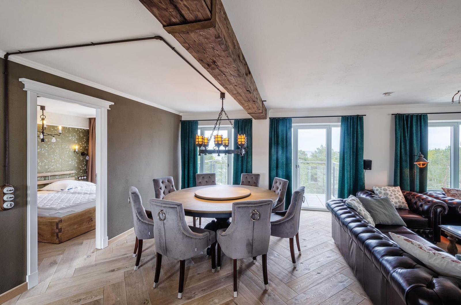 Esszimmer mit runder Tisch und Stühlen, Wohnbereich mit Ledersofa, Blick auf Balkon, Holzbalken an Decke.