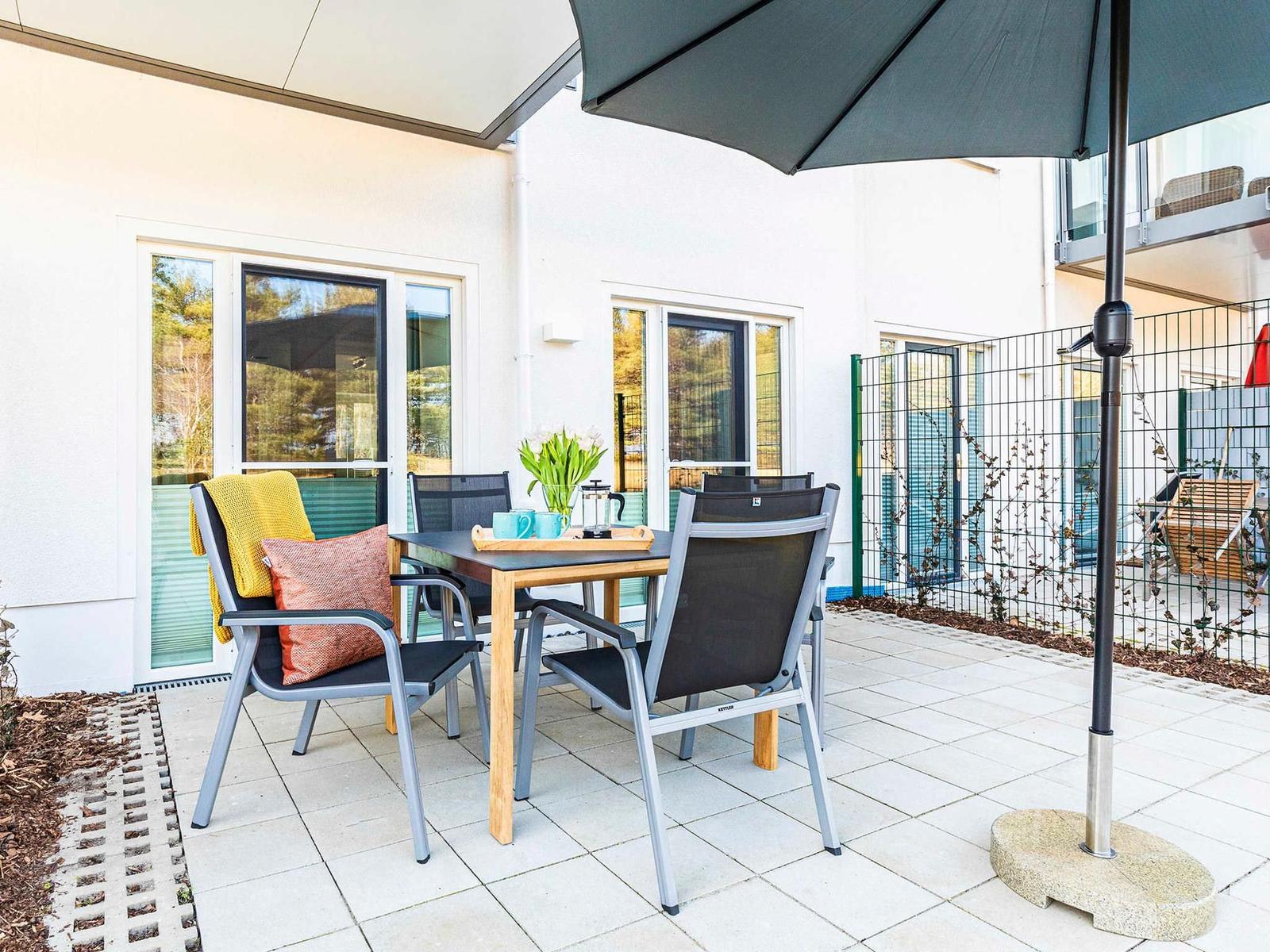 Terrasse mit Tisch, Stühlen und Sonnenschirm. Hintergrund: Haus mit Fenstern und Zaun.