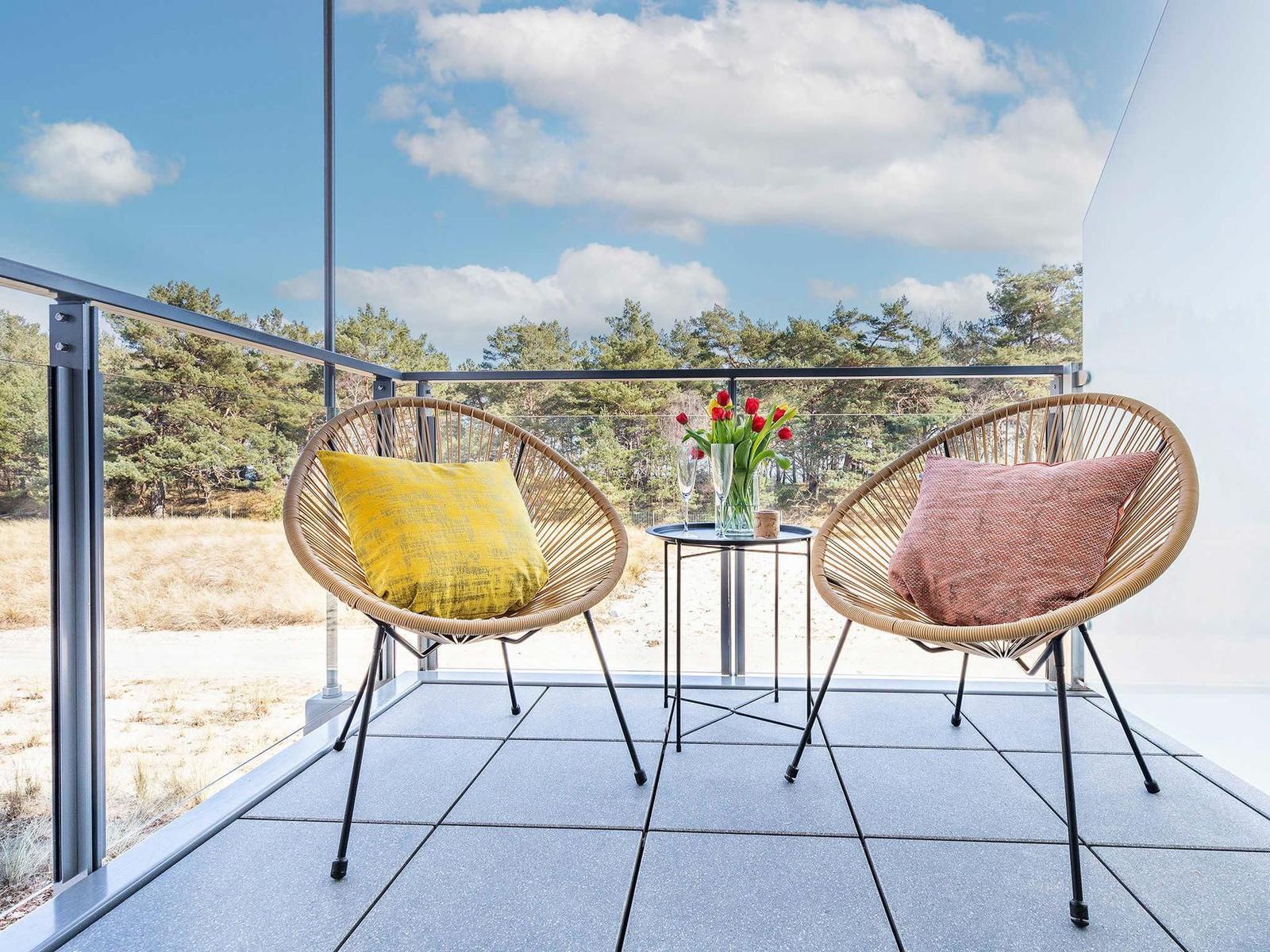 Balcony with two wicker chairs, small table, and tulips. View of pine trees and sky.