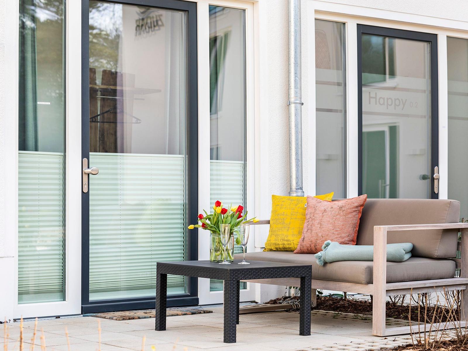 Terrasse mit Sitzbank, Tisch und Blumen vor Glasfront.