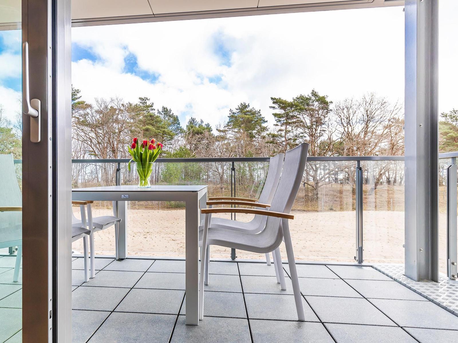 Terrasse mit Tisch, Stühlen und Tulpenblumen. Blick auf Wald und Strand.