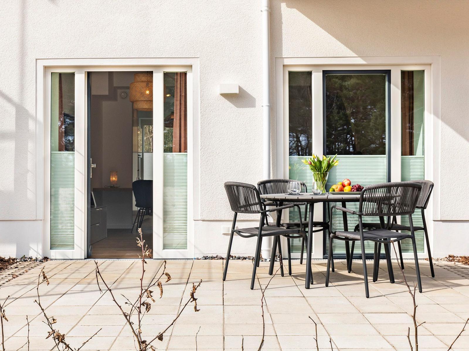Terrasse mit Tisch und Stühlen. Durch Glastüren ist das Innere sichtbar.