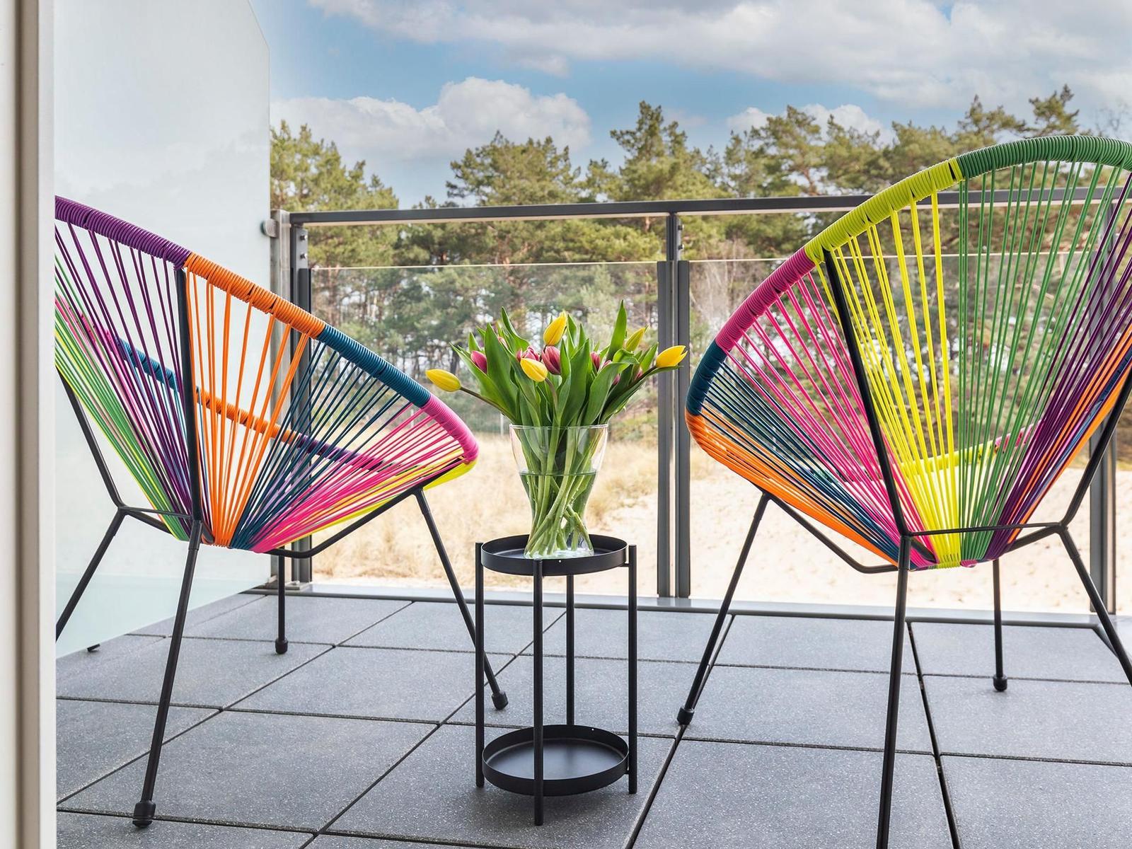 Zwei bunte Sessel und ein Blumenstrauß stehen auf einer Terrasse mit Blick auf einen Wald.