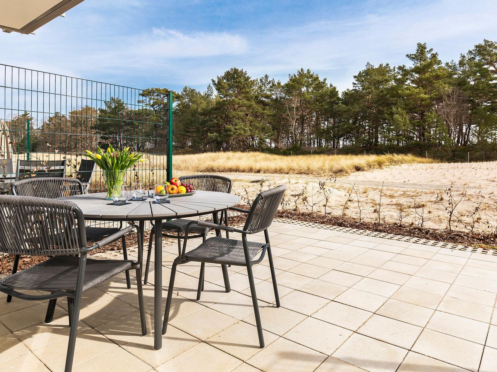 Terrasse mit Tisch und Stühlen, Blick auf Sanddünen und Wald.