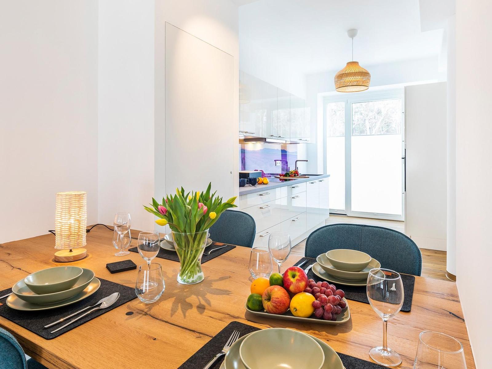 Holztisch mit Geschirr, Blumen und Obst. Küche im Hintergrund mit Fenster.